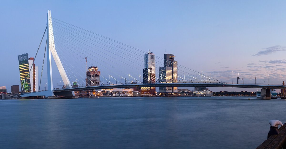 De Erasmusbrug en de skyline van Rotterdam bij avondlicht, gezien vanaf het water.